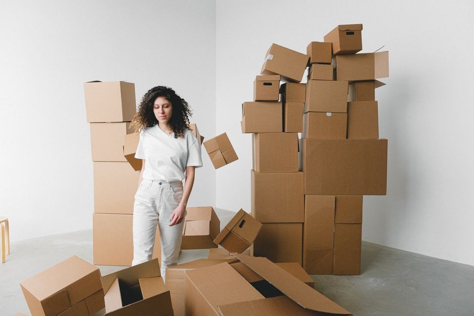 A woman with curly dark hair, dressed in a white t-shirt and light-colored trousers, stands in a room surrounded by numerous flattened cardboard boxes and some upright boxes stacked against the white wall. Several cardboard boxes are on the floor, some open and others sealed, indicating the packing stage of a home relocation. The boxes vary in size and are made of brown corrugated cardboard, with some wrapped in plastic or taped for secure packing. In the background, there is a partially visible wooden stool, and the room has ample natural lighting, contributing to a clean, minimalist environment typical of a professional removals process. This scene depicts the packing, organizing, and preparation phase before loading during a house removal and furniture transport service, consistent with services offered by Man and Van Catford. The setting suggests the interior of a residential property in the process of being prepared for transportation to a new home, emphasizing careful packing and efficient load management.