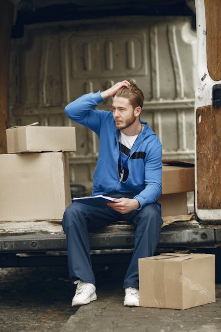 A young man with light brown hair, dressed in a blue sweatshirt, navy trousers, and white sneakers, is sitting inside the open back of a light-colored van parked on a street. He is holding a clipboard